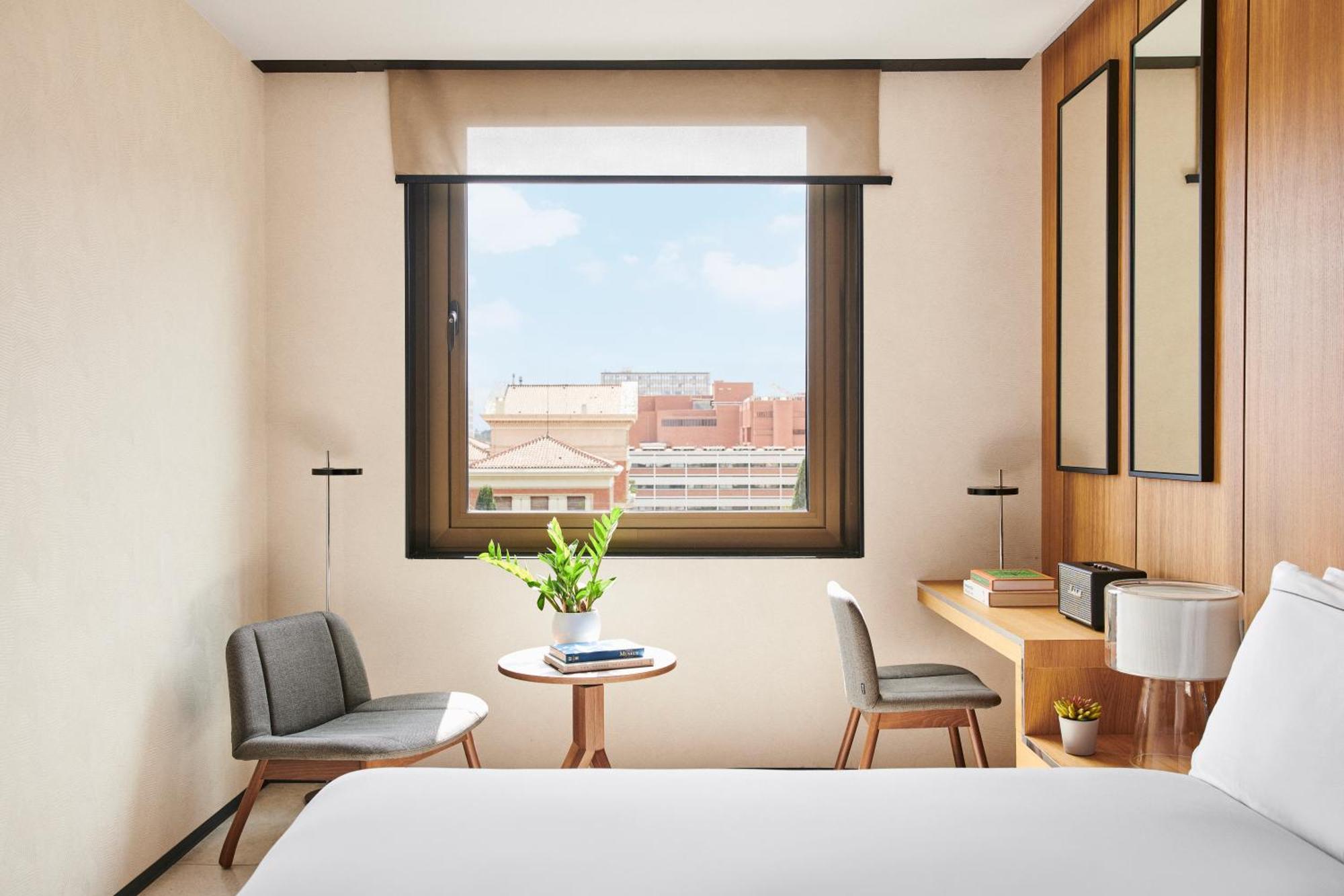 Hotel Grand Hyatt Barcelona Exteriér fotografie The photo depicts a modern hotel room with a cozy and minimalist design. There's a large window allowing natural light to enter, showcasing a view of the outside cityscape. In the room, two chairs are positioned near a small round table, which has a 