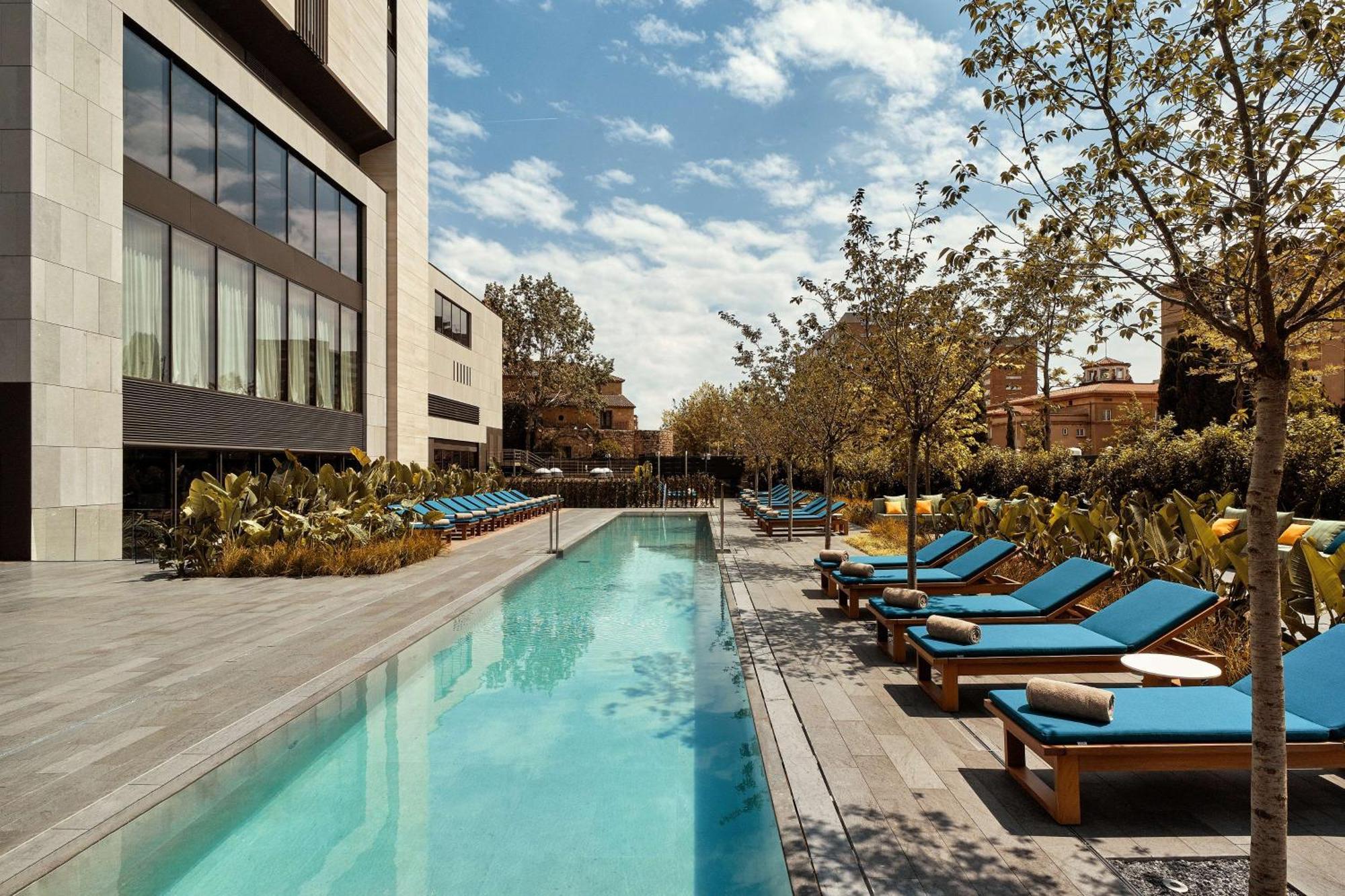 Hotel Grand Hyatt Barcelona Exteriér fotografie The photo showcases a modern outdoor pool area. In the foreground, there is a long, rectangular swimming pool with clear blue water. Surrounding the pool are lounge chairs with bright blue cushions, arranged neatly along both sides. The area features